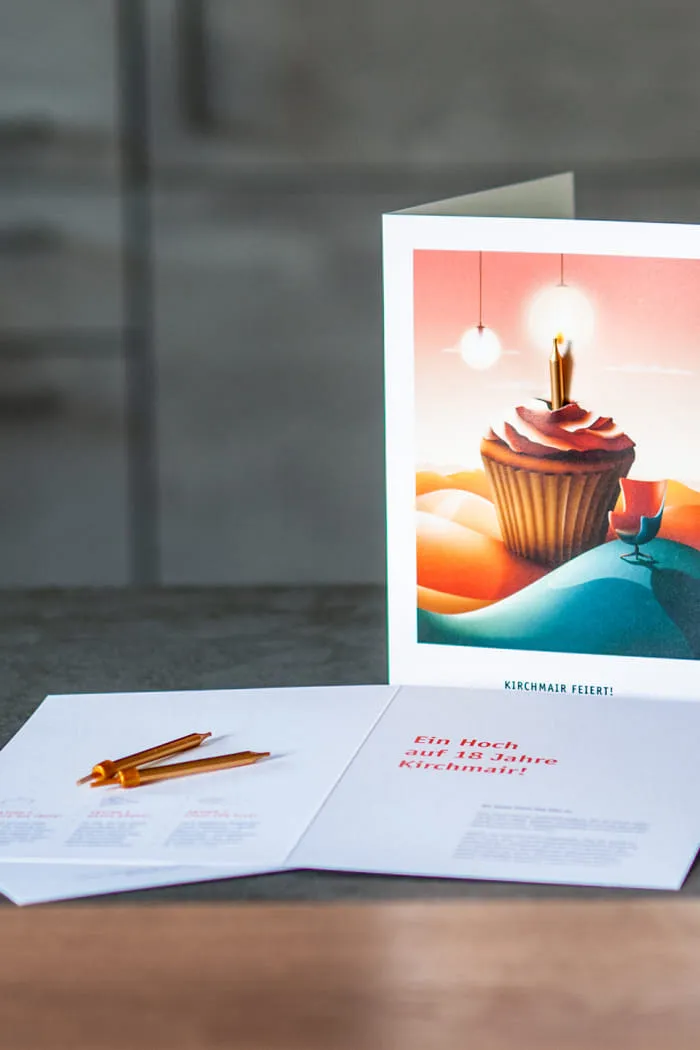 Auf grauer Stein- und Holzplatte steht eine aufgeschlagenen Grußkarte. Die Vorderseite zeigt eine Illustration mit einem großen Cupcake, einer Kerze und surrealen Elementen wie Hügeln, Leuchten und einem Designersessel. Daneben liegt eine geöffnete Grußkarte mit Text zum 18-jährigen Bestehen von Kirchmair und 2 goldenen Kerzen.