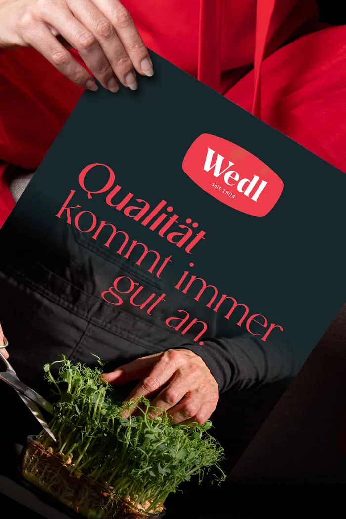 Eine Hand hält die Anzeige im neuen Wedl-Design mit dem neuen Logo. Dahinter ist ein roter Hintergrund zu sehen. Das Design zeigt das Wedl-Logo, Kresse, eine Hand und den Claim der Kampagne.