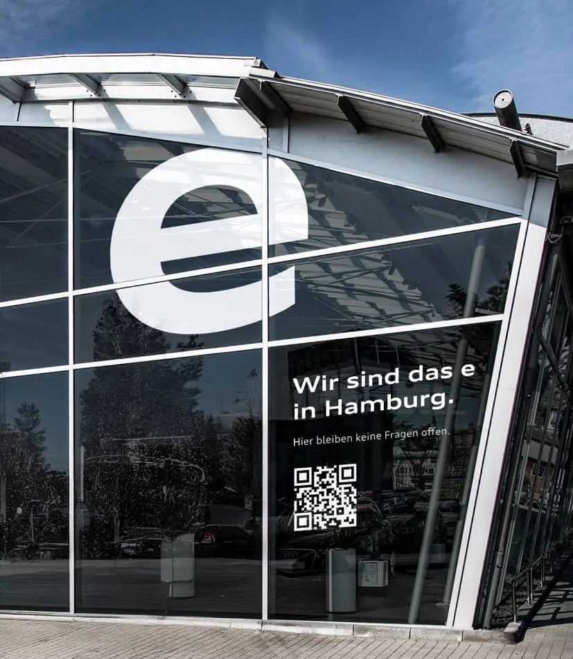 Die Glasfassade eines Audi-Autohauses in Hamburg mit großem weißen e-Logo und dem Kampagnenslogan: Wir sind das e in Hamburg sowie dem Zusatz: Hier bleiben keine Fragen offen, und einem QR-Code darunter.