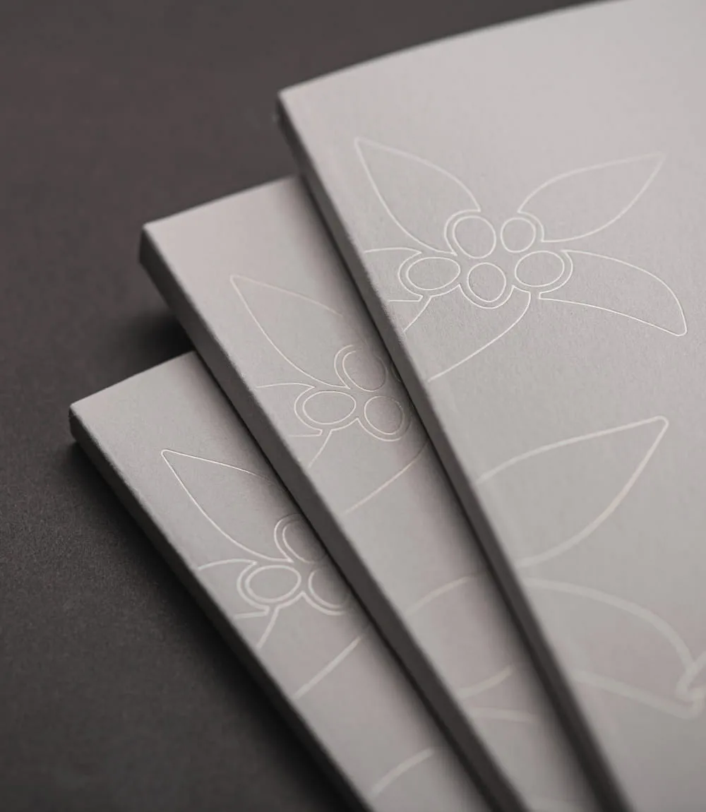Nahaufnahme gestaffelter weißer Hotelbroschüren mit elegant geprägtem Blumenmuster. Die dezente Relieftechnik zeigt stilisierte Alpenblüten auf hochwertigem Papier.