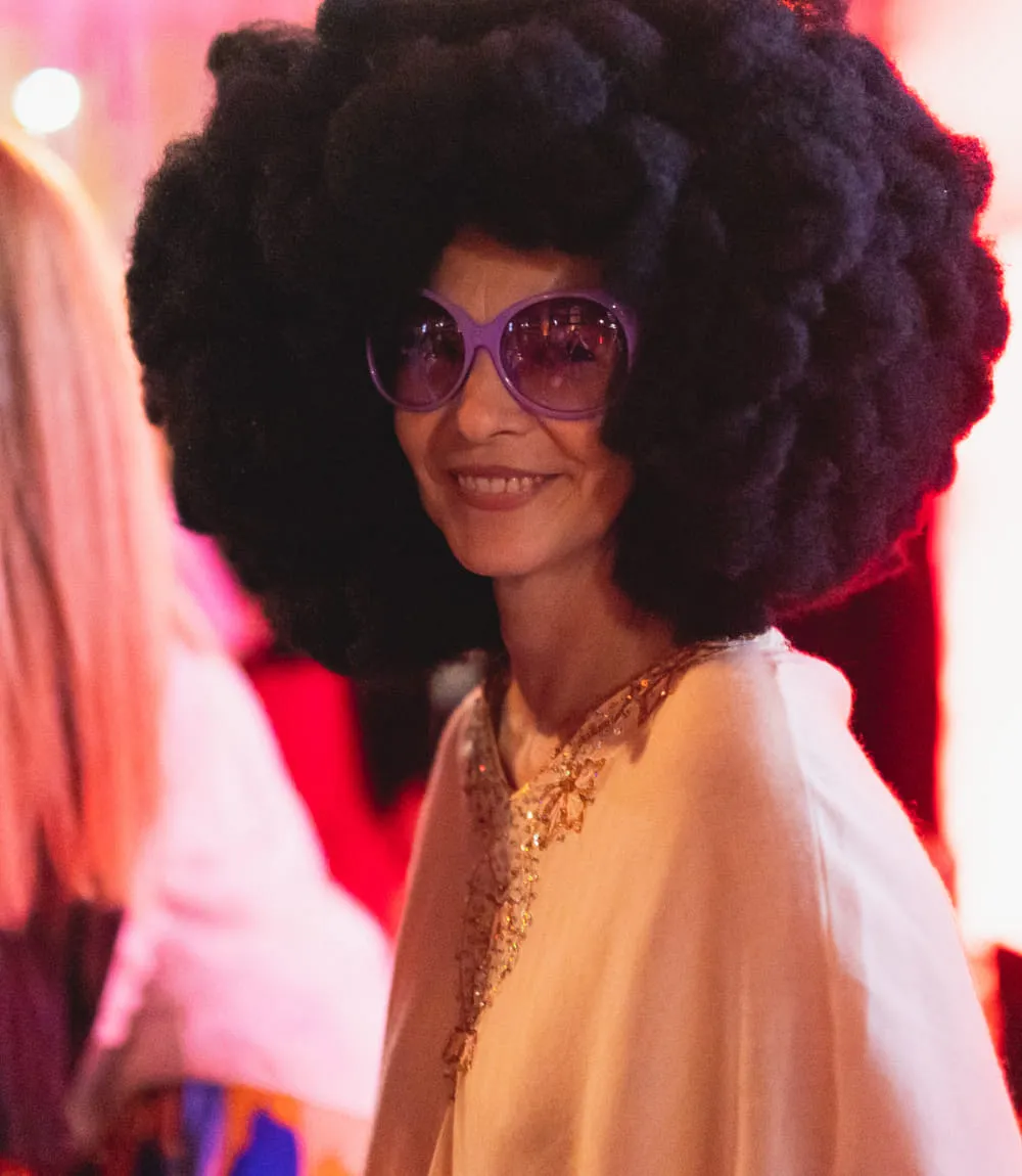 Frau mit großer Afro-Perücke und lila Sonnenbrille lächelt in die Kamera. 