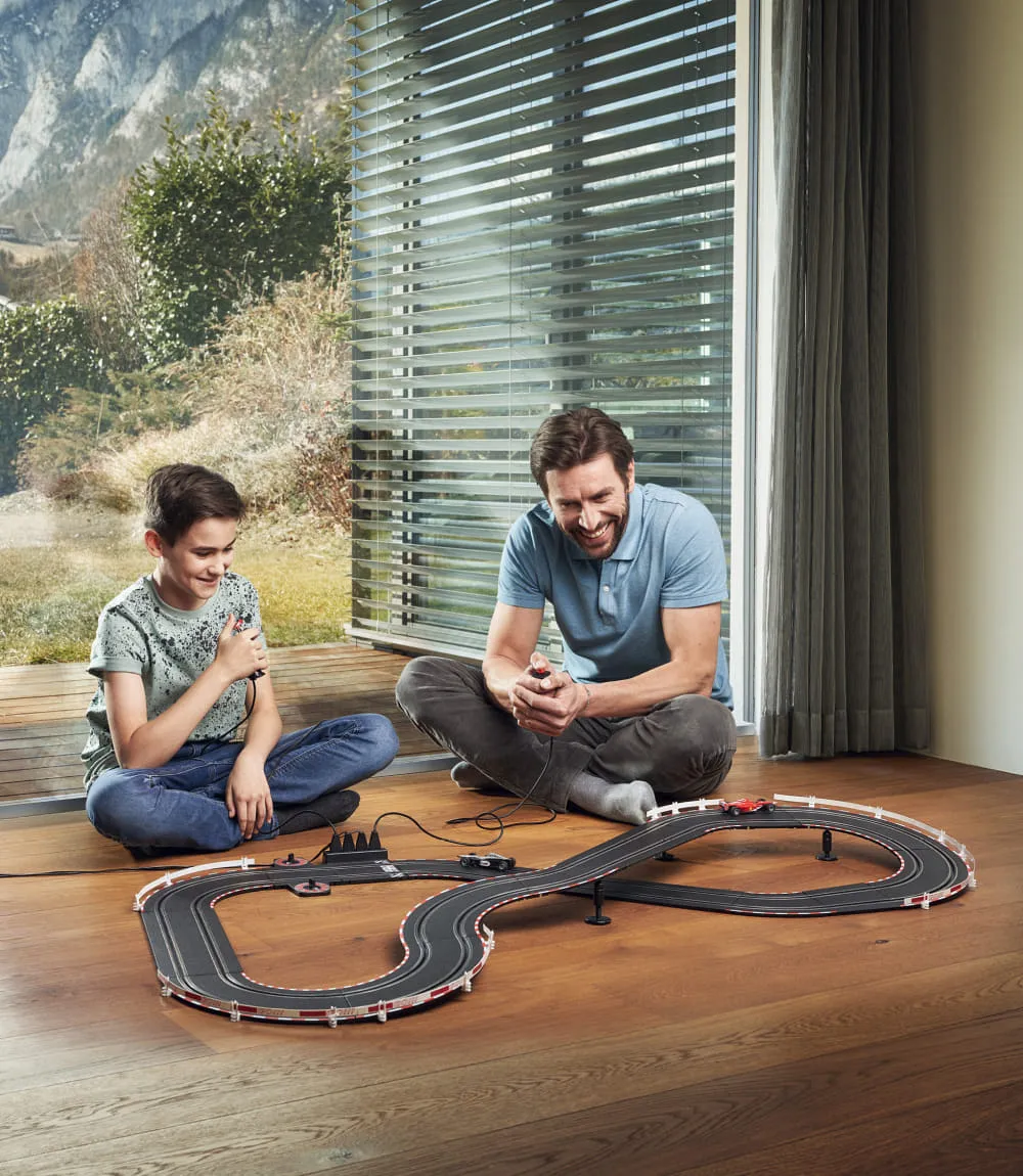 Vater und Sohn spielen gemeinsam mit einer Carrera-Rennbahn auf einem Holzboden vor einem großen Fenster mit Bergpanorama.