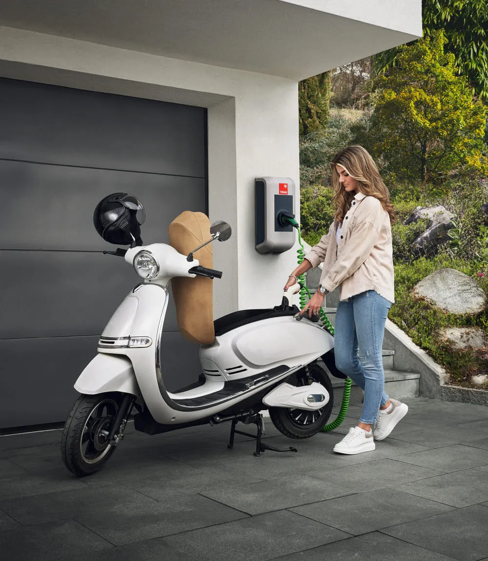 Junge Frau lädt einen weißen Elektroroller an einer TIWAG-Wandladestation vor einer modernen Garage mit grünem Naturgarten im Hintergrund.