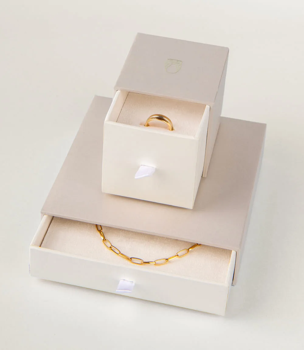 Zwei Schmuckverpackungen in Creme und Beige, eine mit goldener Halskette, die andere mit einem Ring.