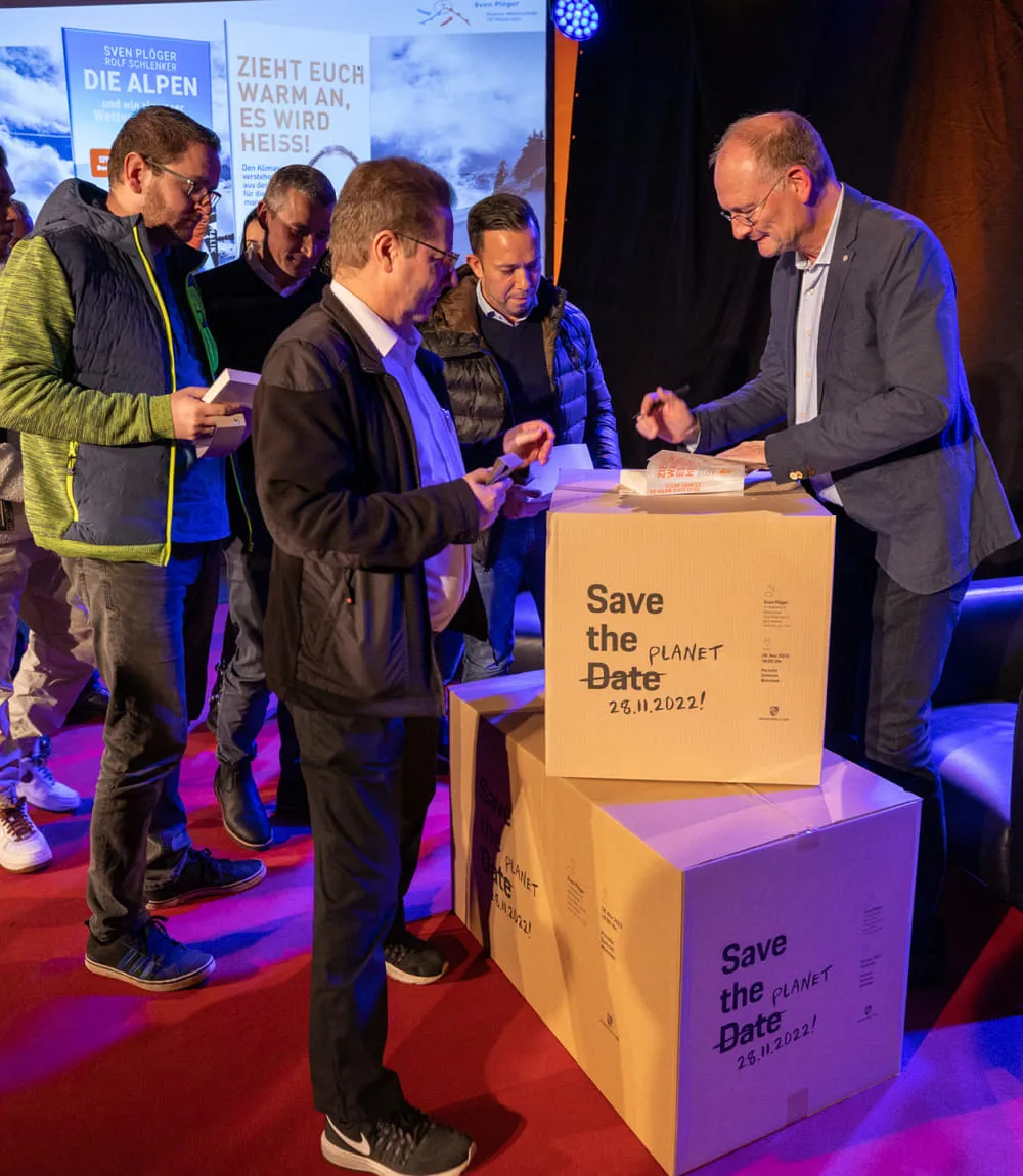 Gruppe von Männern steht für Büchersigning von Sven Plöger an. Er benutzt als Unterlage übereinandergespapelte Kartons die das Motto Save the Date - Save the Planet präsentieren.